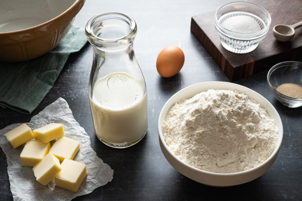 the ingredients to make Danish brunsviger cake: a bowl of flour, pads of butter on parchment paper, active dry yeast in a glass bowl, an egg, and granulated sugar in a ramekin