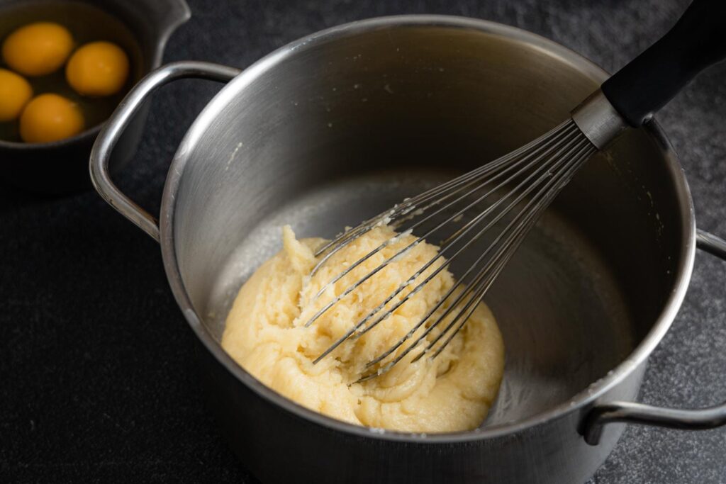 choux paste in a saucepan with a whisk and eggs in a prep bowl